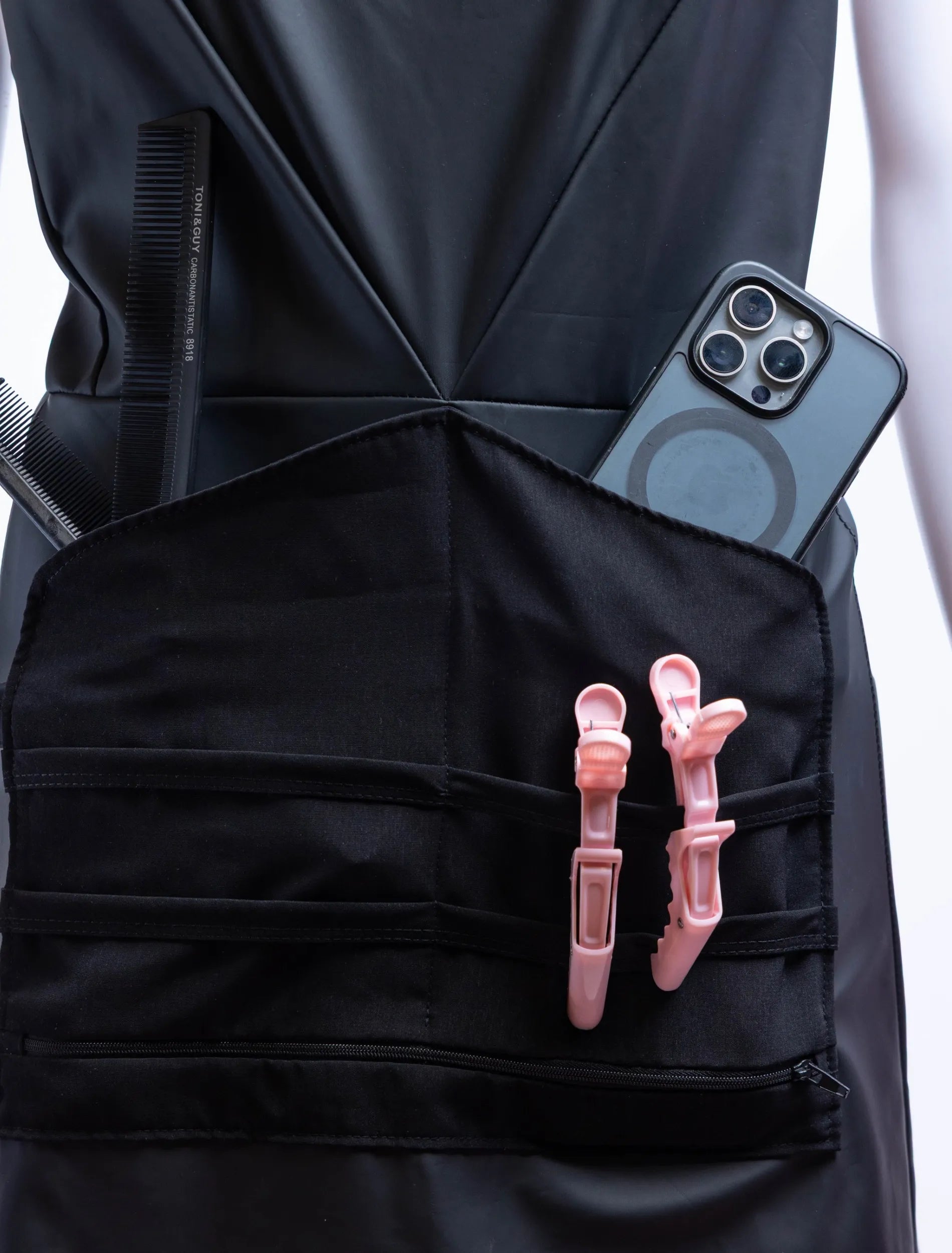 A detailed front-view photograph of a black stylist apron from The Branded Salon on a mannequin, showcasing the professional tool organizer panel holding black TONI&GUY combs, a smartphone, and light pink hair clips, with a heart-shaped metal ring on the side.