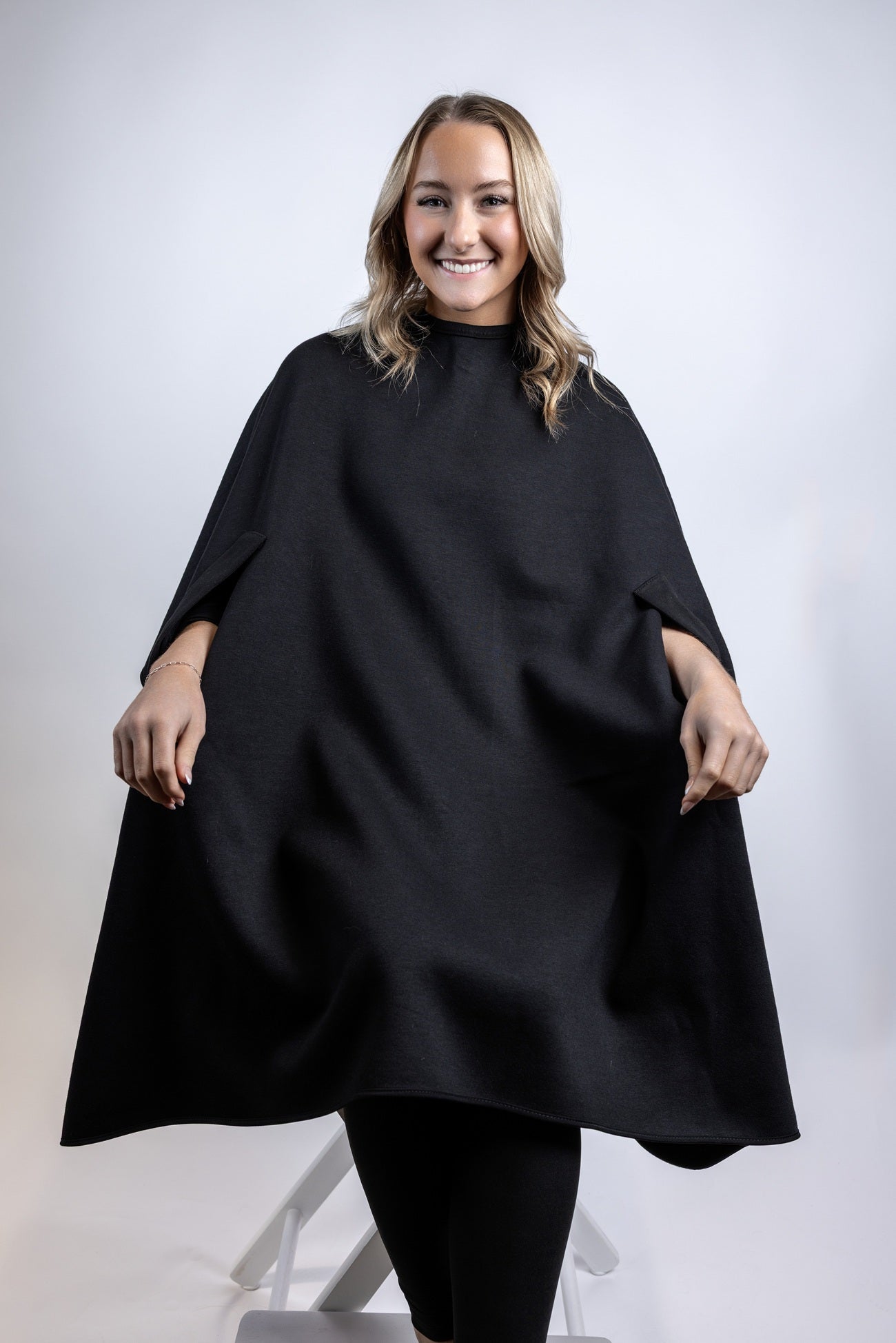 A portrait of a woman smiling at the camera while wearing a black salon cape featuring integrated arm slits for client comfort, set against a plain light background.