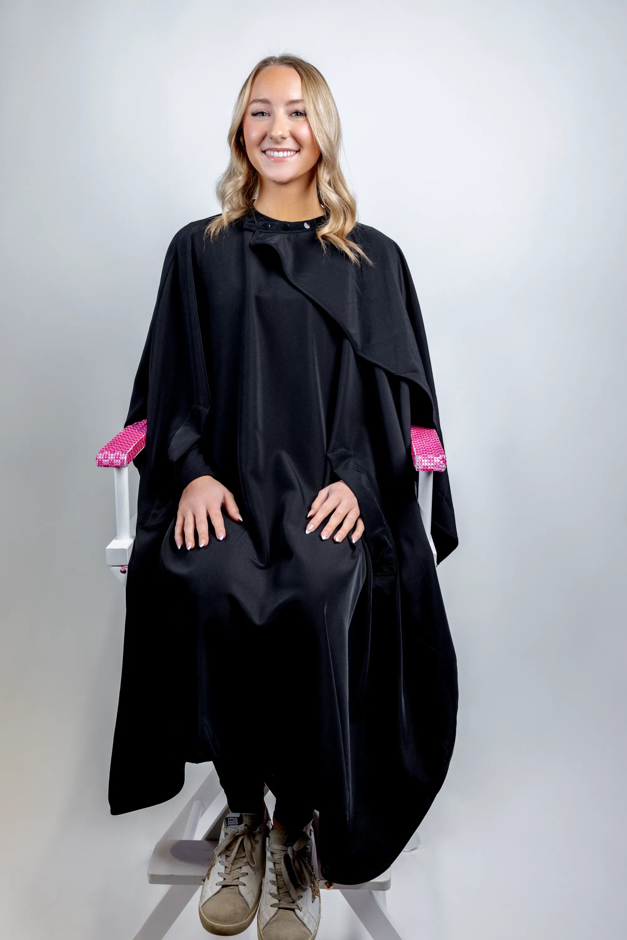 A full-length lifestyle photograph of a smiling woman seated in a white director's chair with pink rhinestone armrests, wearing a professional, full-coverage black salon cape from The Branded Salon.