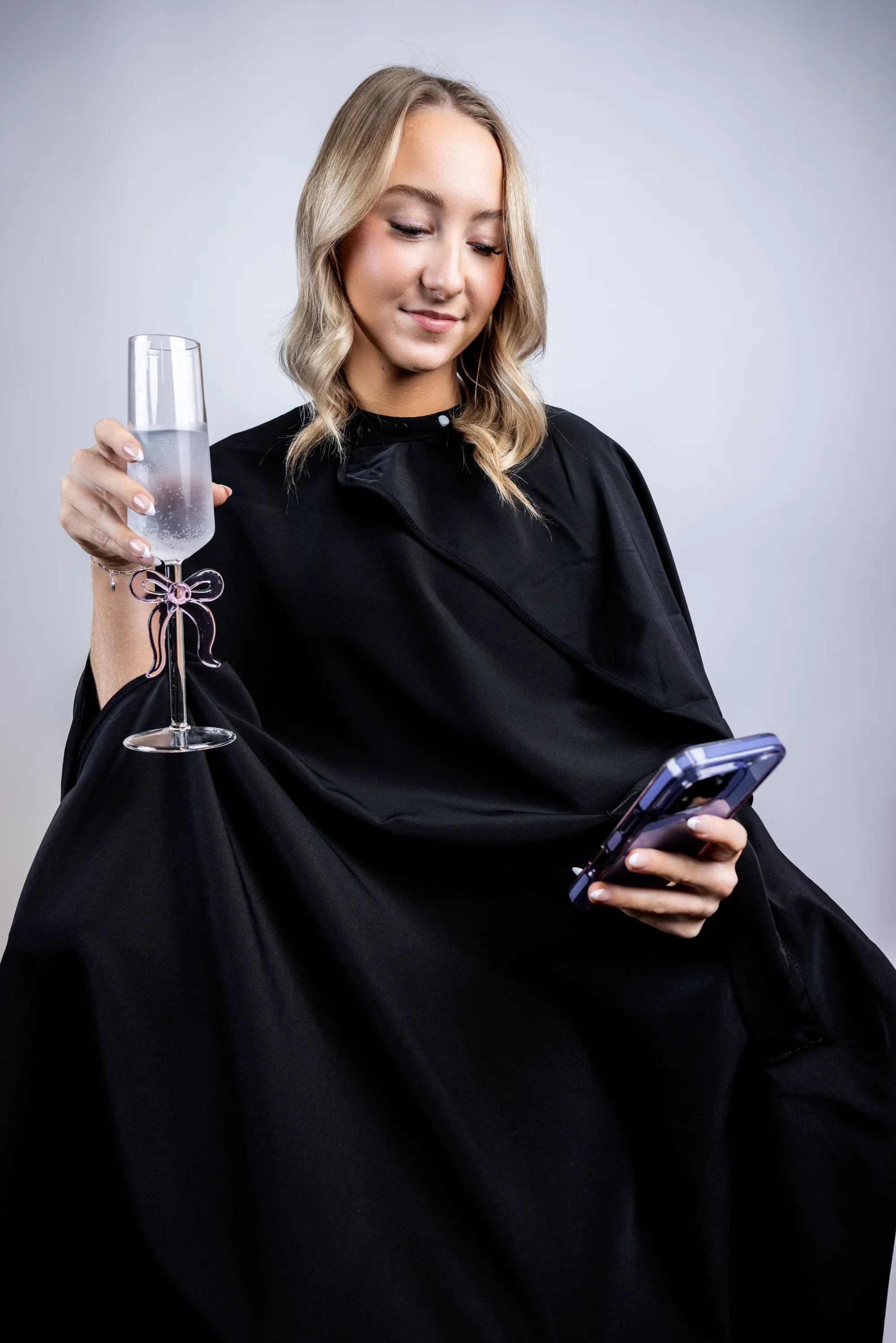 A lifestyle photograph of a woman wearing a professional black salon cape, multi-tasking by holding a champagne flute with a pink bow and looking at her smartphone while seated.