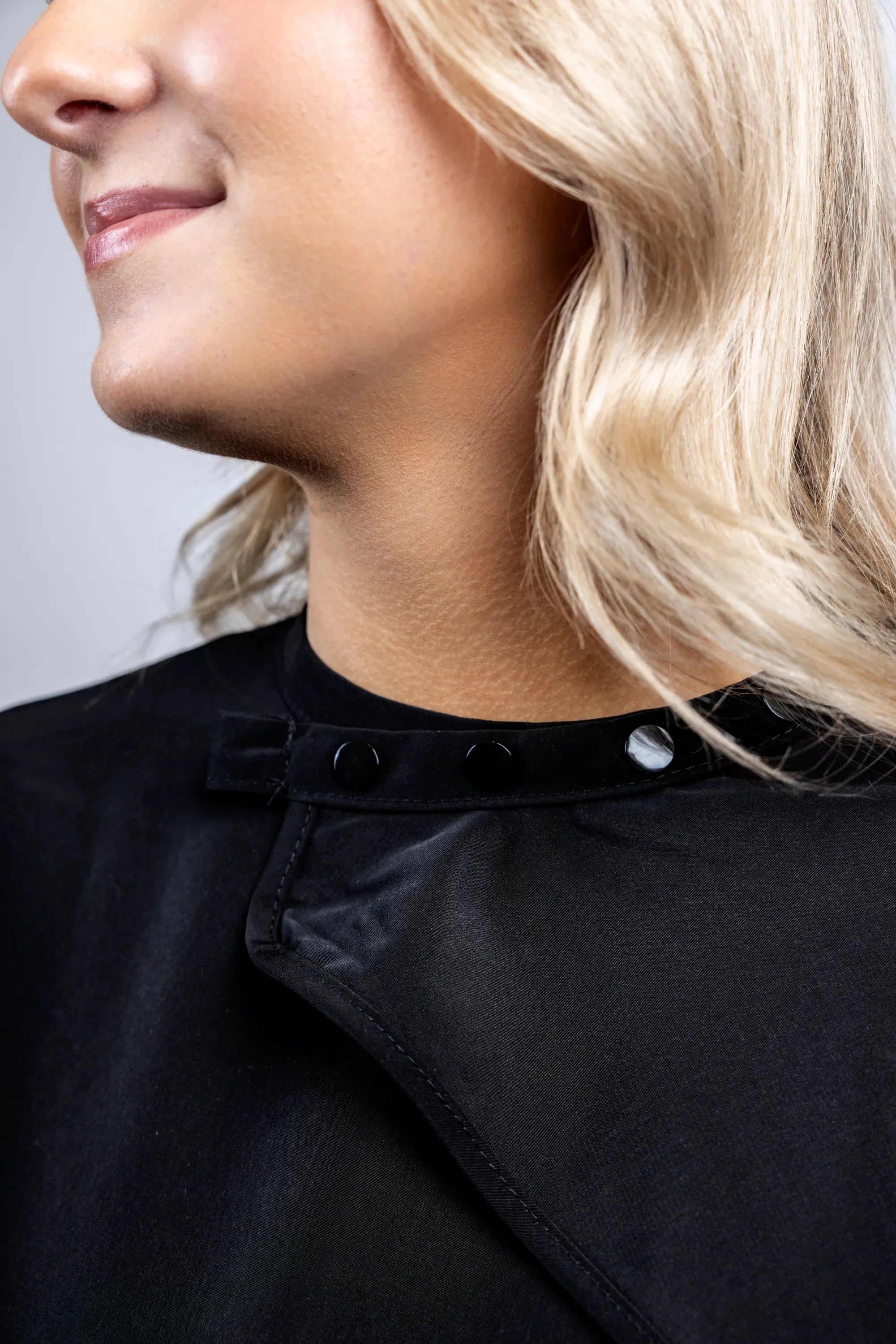 A close-up detail shot focusing on the black snap-button neck closure of a professional black salon cape, showing the secure and adjustable fasteners.