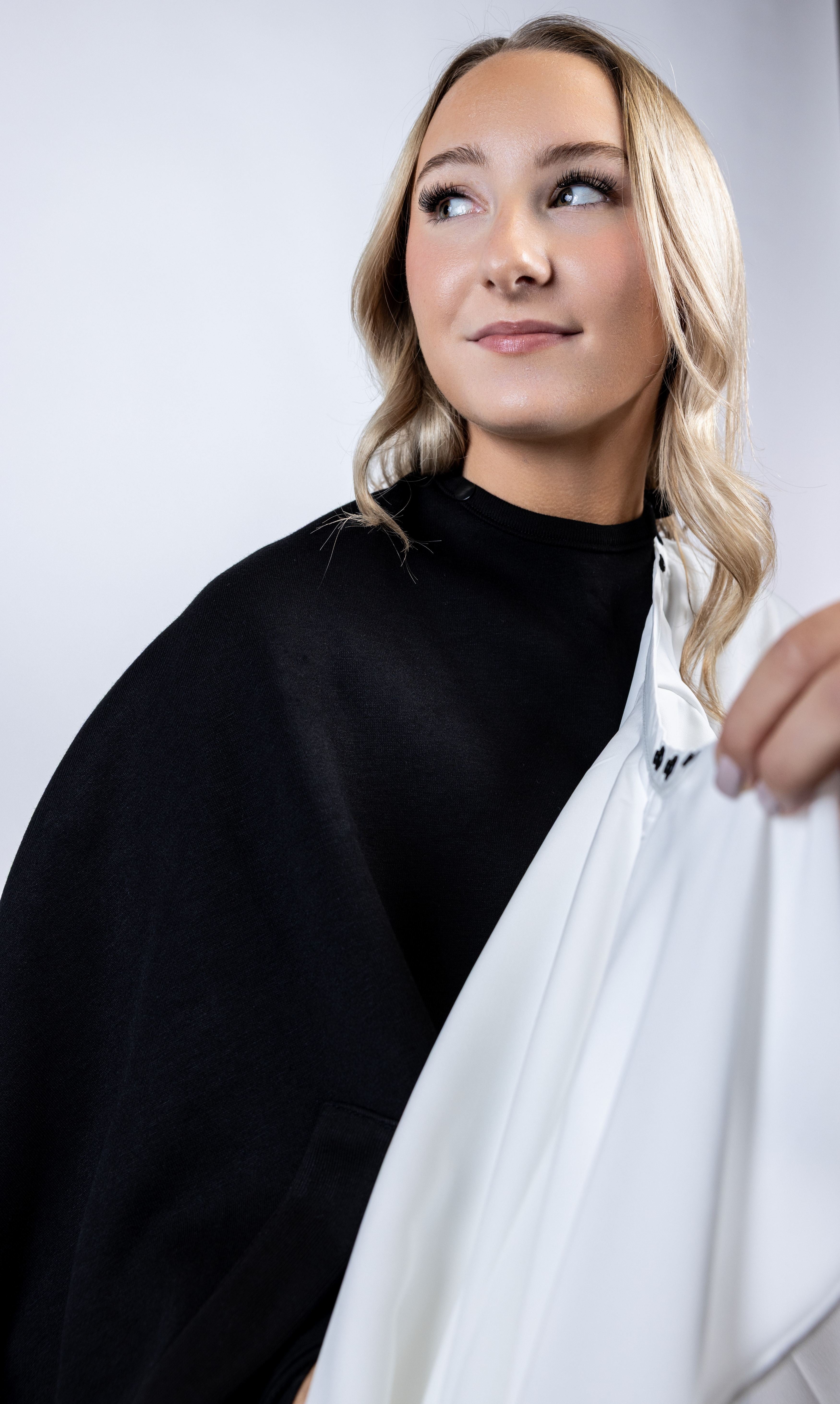 A close-up lifestyle photograph of a woman wearing a black salon cape while a white salon cape is held beside it to show the different color options available in the apparel line.