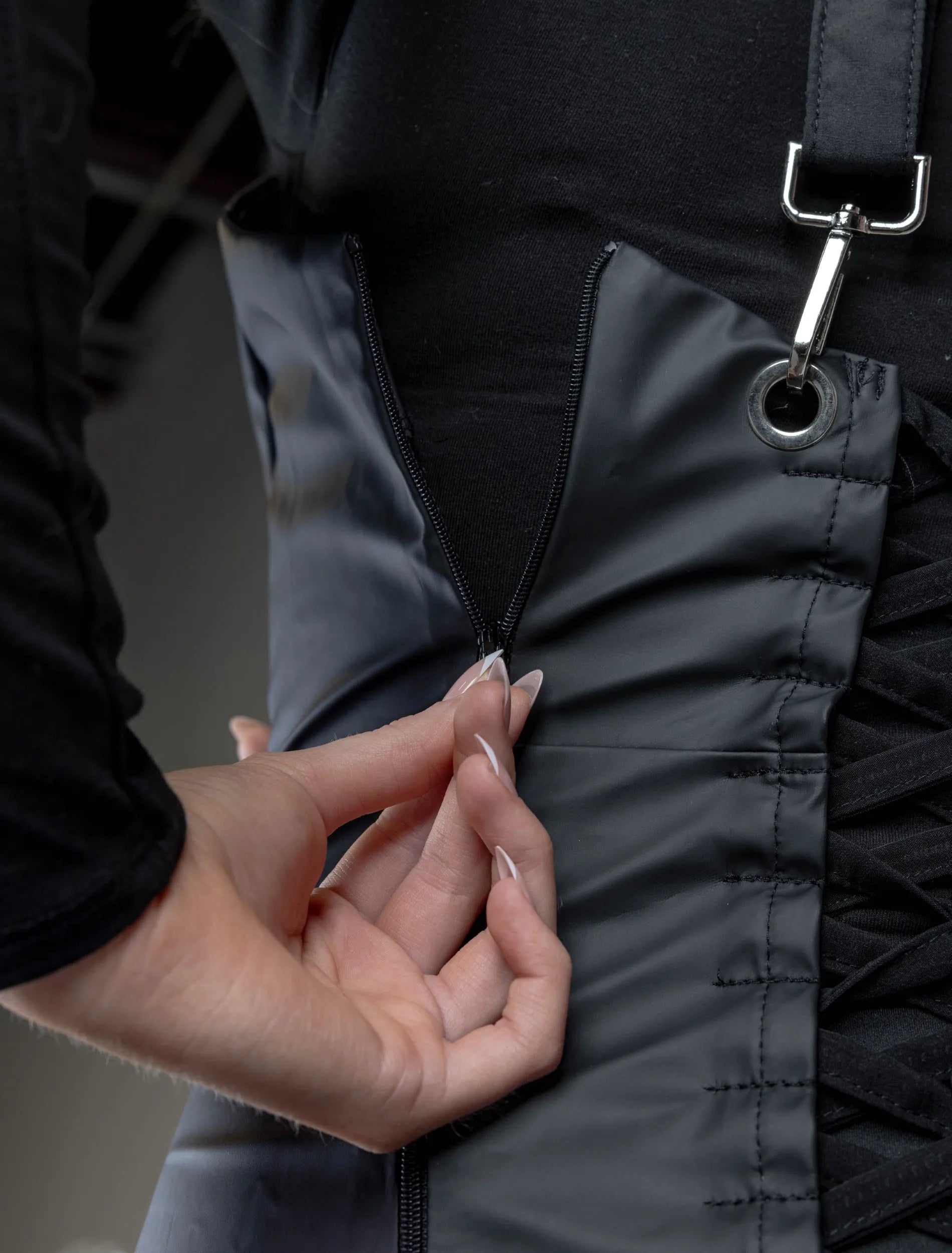 A close-up detail photograph of the back of a black stylist apron from The Branded Salon, showing a hand with French-tipped nails operating a zipper next to a corset-style lace-up closure and metal eyelets.
