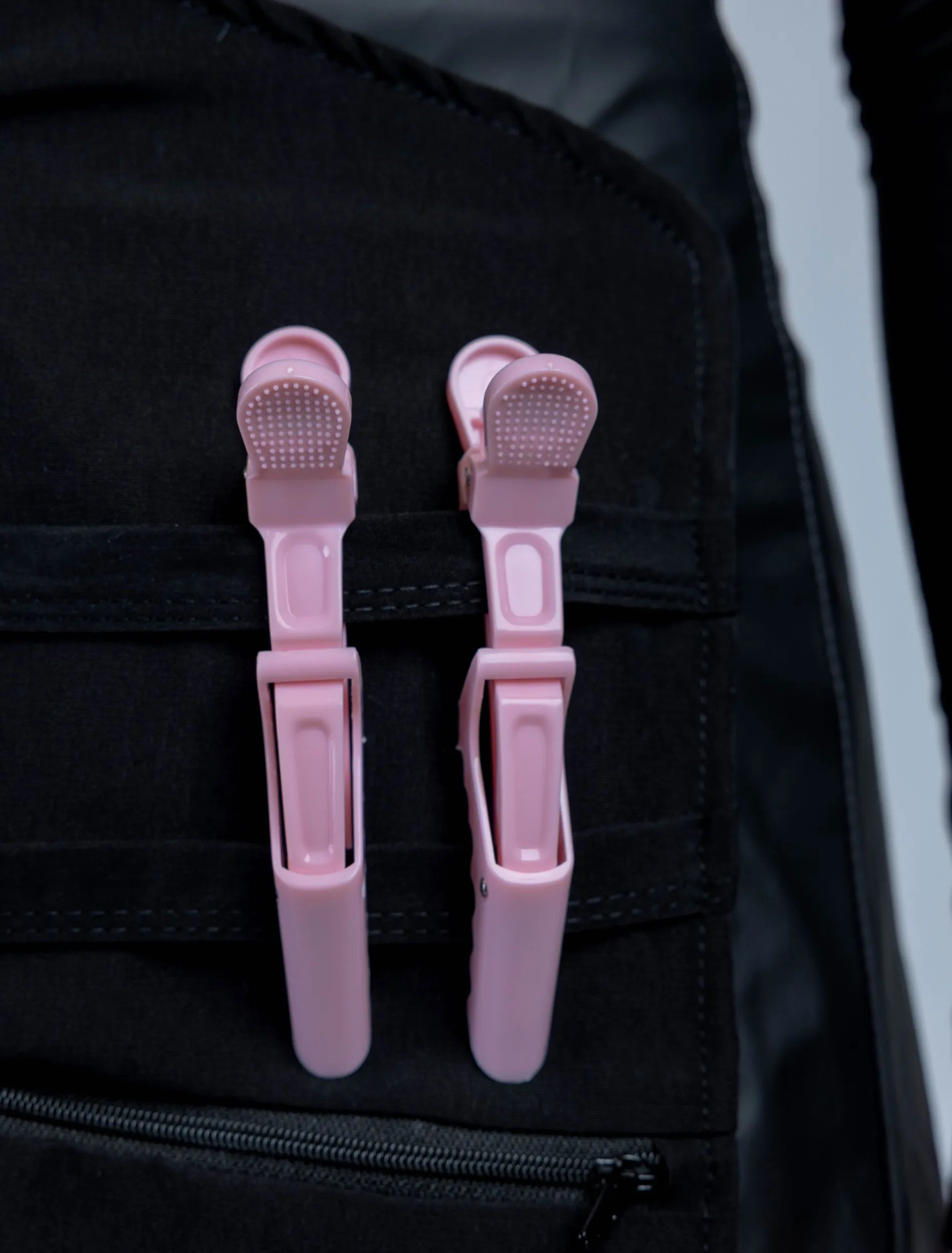 A close-up detail photograph of a black stylist apron from The Branded Salon, featuring two light pink hair clips attached to the horizontal straps of a specialized tool organizer panel.