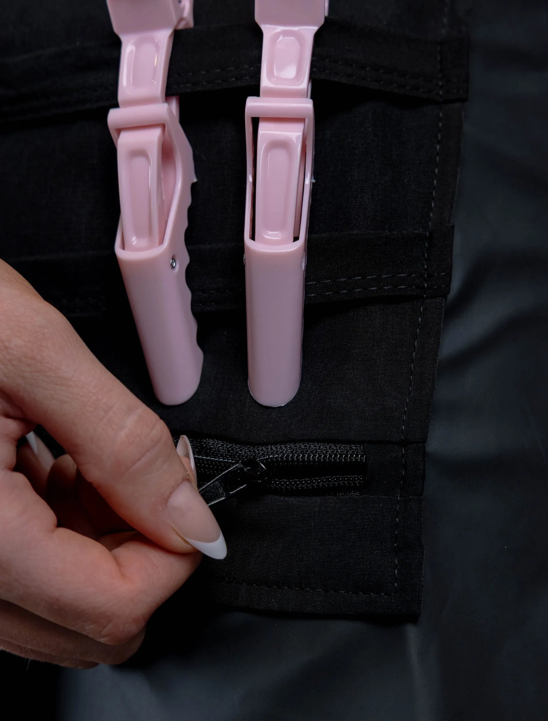A close-up detail photograph of the tool organizer panel on a black stylist apron, featuring two pink hair clips and a hand with French-tipped nails operating a black zipper closure at the bottom.