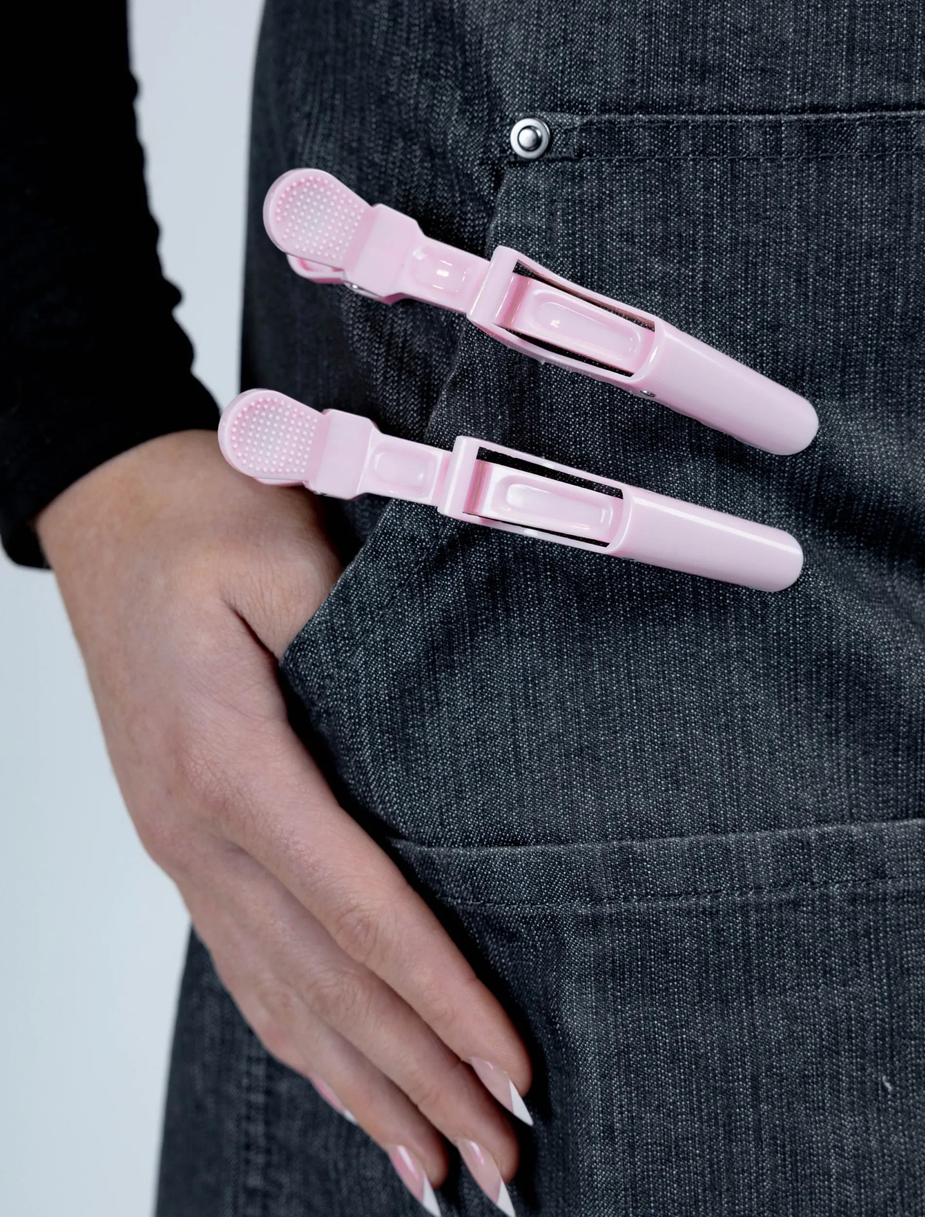 A close-up detail photograph of a hand with French-tipped nails tucked into the pocket of a charcoal grey denim stylist apron, featuring two light pink hair clips attached to the pocket's edge.