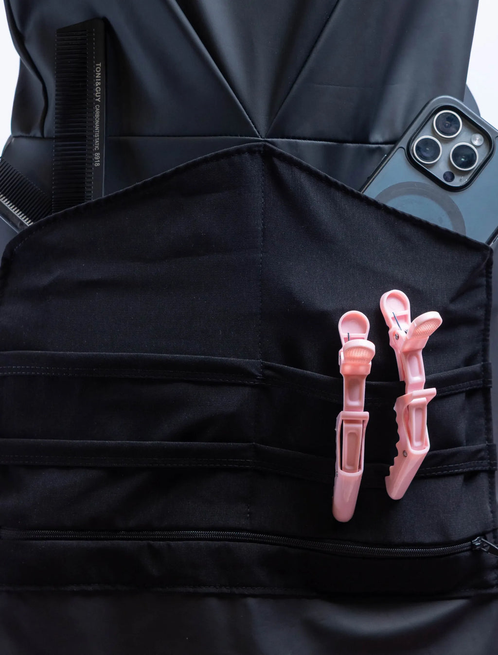 A front-view photograph of a black professional stylist apron from The Branded Salon displayed on a mannequin, showcasing the central tool organizer panel holding black combs, a smartphone, and two light pink hair clips, with a silver heart-shaped ring visible on the side.