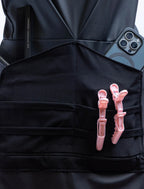 A front-view photograph of a black professional stylist apron from The Branded Salon displayed on a mannequin, showcasing the central tool organizer panel holding black combs, a smartphone, and two light pink hair clips, with a silver heart-shaped ring visible on the side.