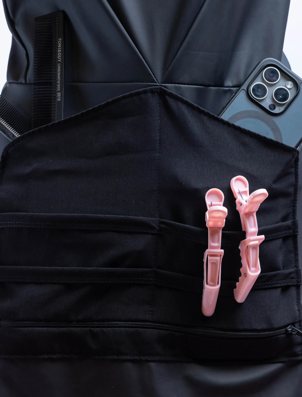 A front-view photograph of a black professional stylist apron from The Branded Salon displayed on a mannequin, showcasing the central tool organizer panel holding black combs, a smartphone, and two light pink hair clips, with a silver heart-shaped ring visible on the side.