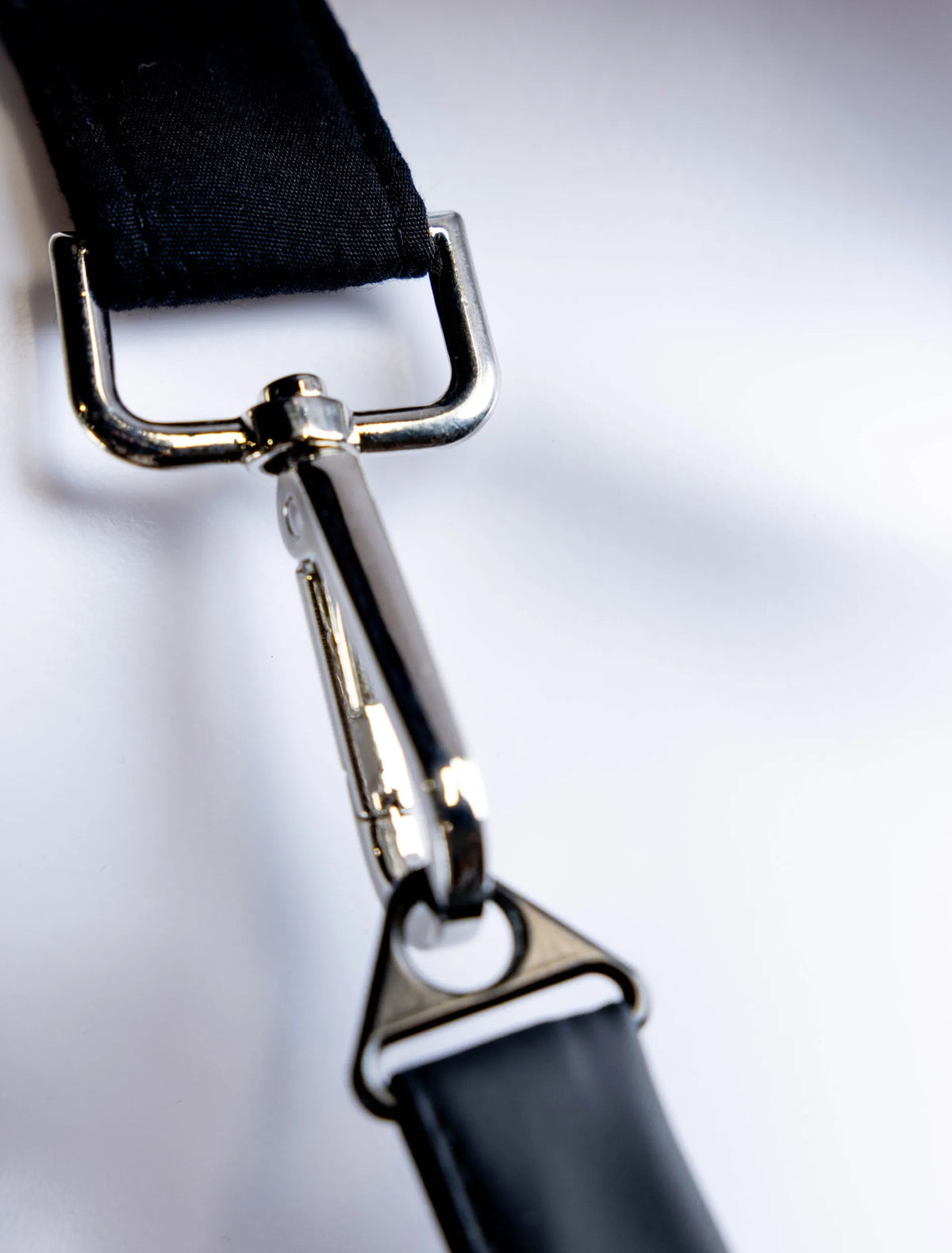 A close-up detail photograph of professional silver metal hardware, specifically a swivel snap hook, attaching a black shoulder strap to the body of a stylist apron from The Branded Salon, displayed on a white mannequin.