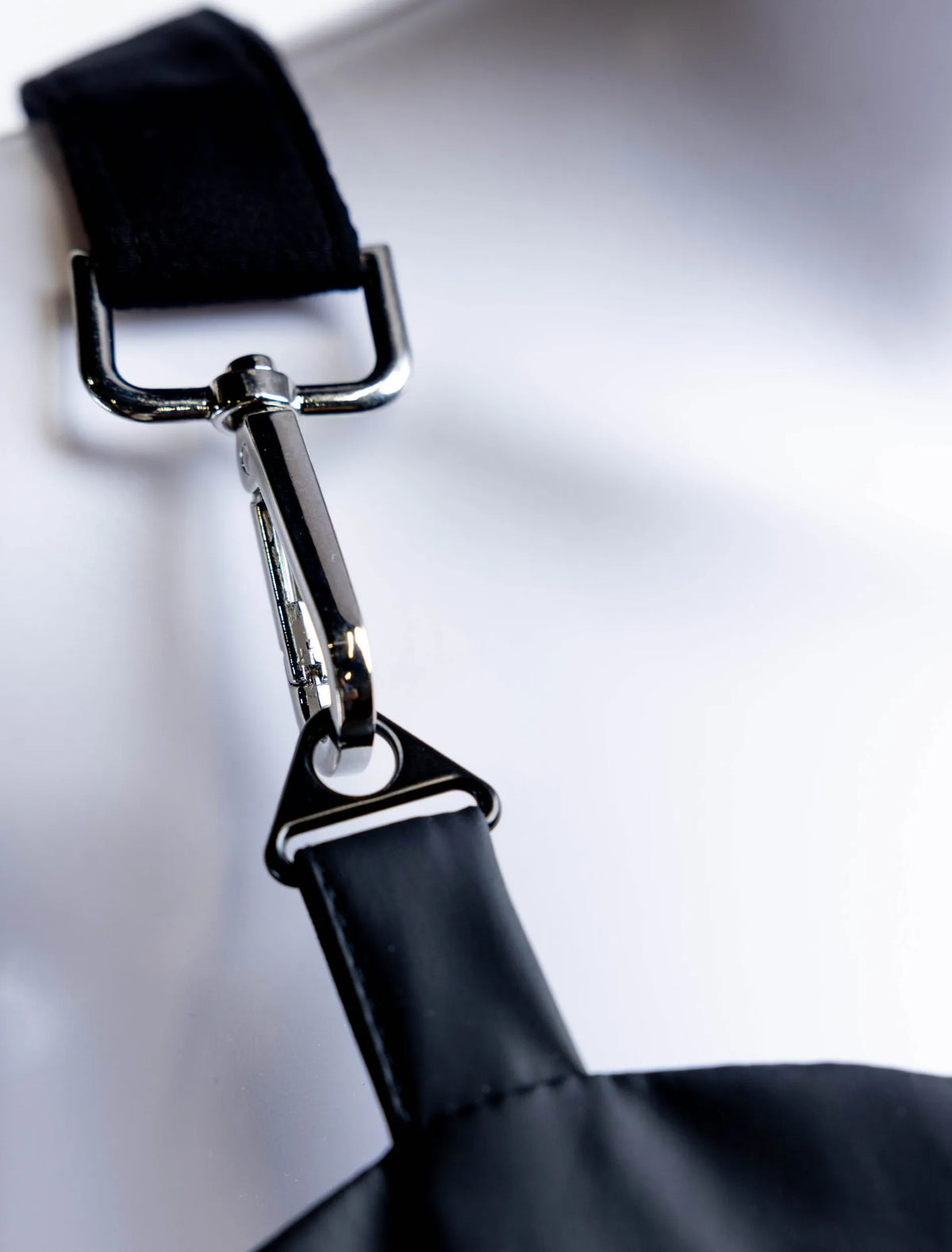 A close-up detail photograph of a silver metal swivel snap hook hardware component that attaches a black shoulder strap to the top edge of a professional stylist apron from The Branded Salon, shown on a white mannequin.