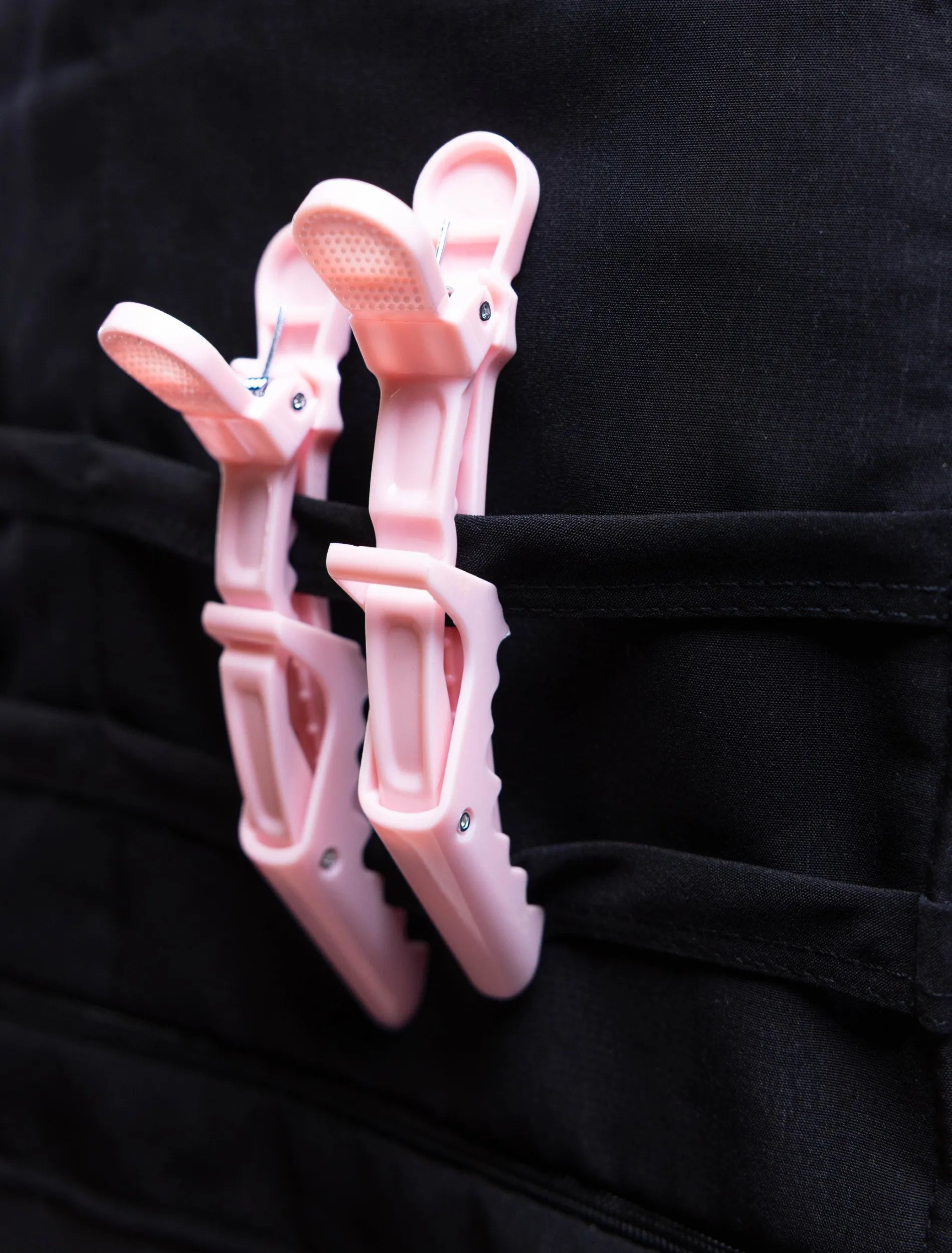 A close-up detail photograph of two light pink professional hair clips attached to a horizontal strap on the tool organizer panel of a black stylist apron from The Branded Salon.