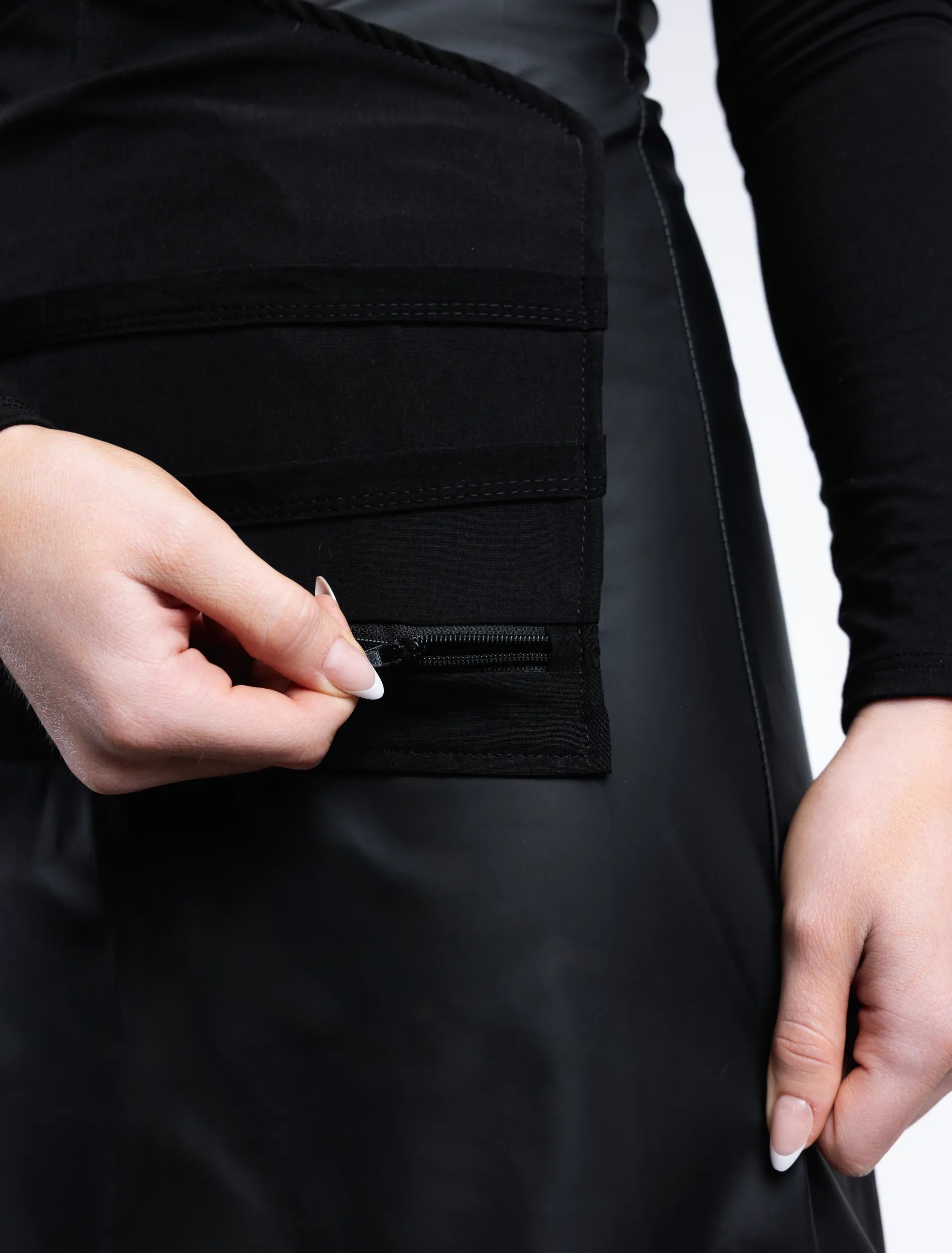 A close-up detail photograph focusing on a hand with French-tipped nails operating a black zipper closure at the base of the specialized tool organizer panel on a black stylist apron from The Branded Salon.