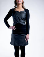 A full front-view photograph of a blonde woman wearing a black professional stylist apron from The Branded Salon, featuring a sweetheart neckline and a specialized central tool organizer panel.