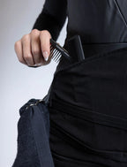 A close-up detail photograph of a black stylist apron from The Branded Salon, featuring a hand with French-tipped nails placing a black TONI&GUY carbon antistatic comb into a pocket on the central tool organizer panel, next to a metal ring with a professional towel attached.

