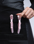 A close-up detail photograph of a hand with French-tipped nails adjusting light pink hair clips on the specialized tool organizer panel of a black professional stylist apron.