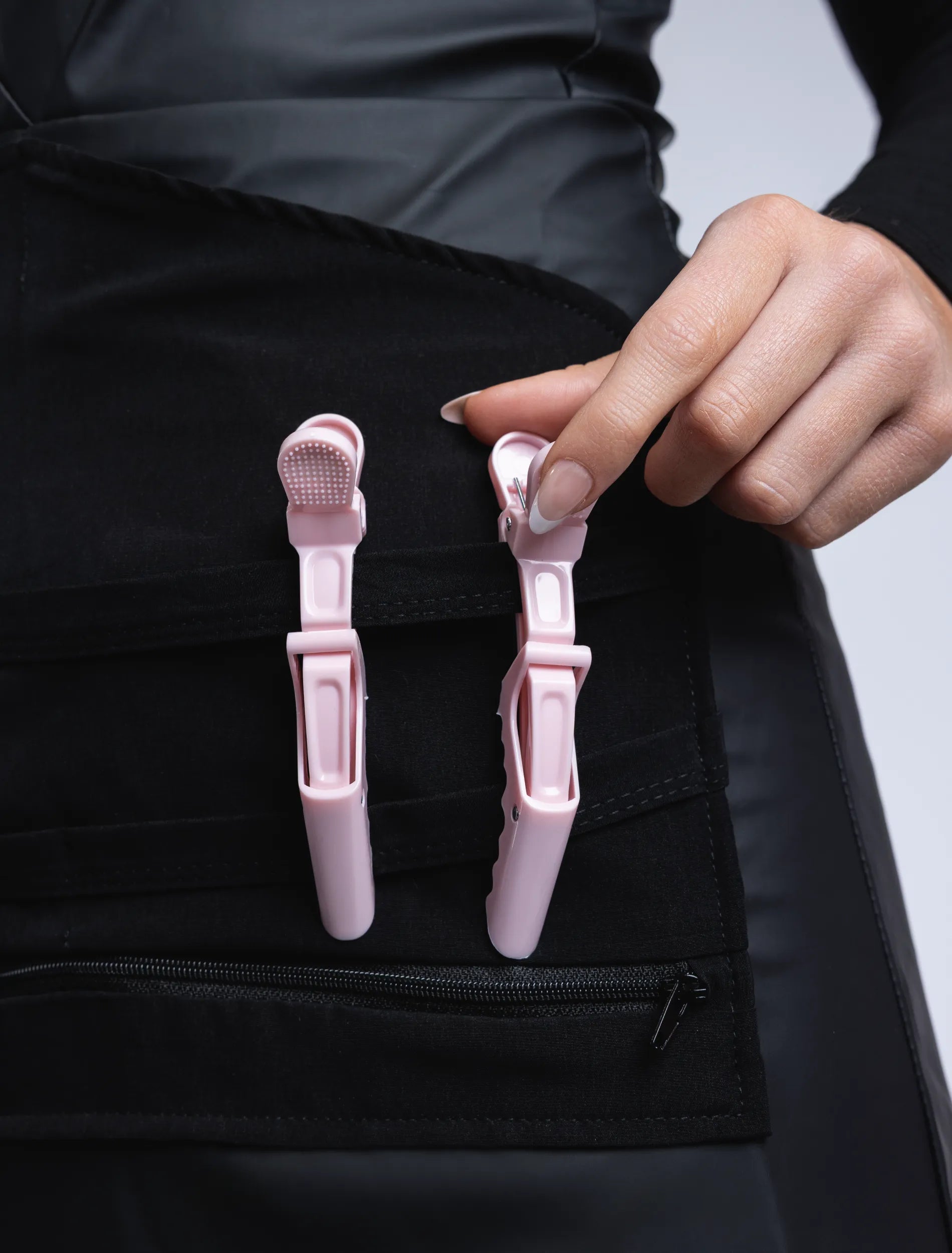 A close-up detail photograph of a hand with French-tipped nails adjusting light pink hair clips on the specialized tool organizer panel of a black professional stylist apron.