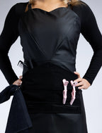 A medium front-view photograph of a black sweetheart neckline stylist apron from The Branded Salon, showcasing the central tool panel with pink hair clips, professional combs, and a towel ring attachment.