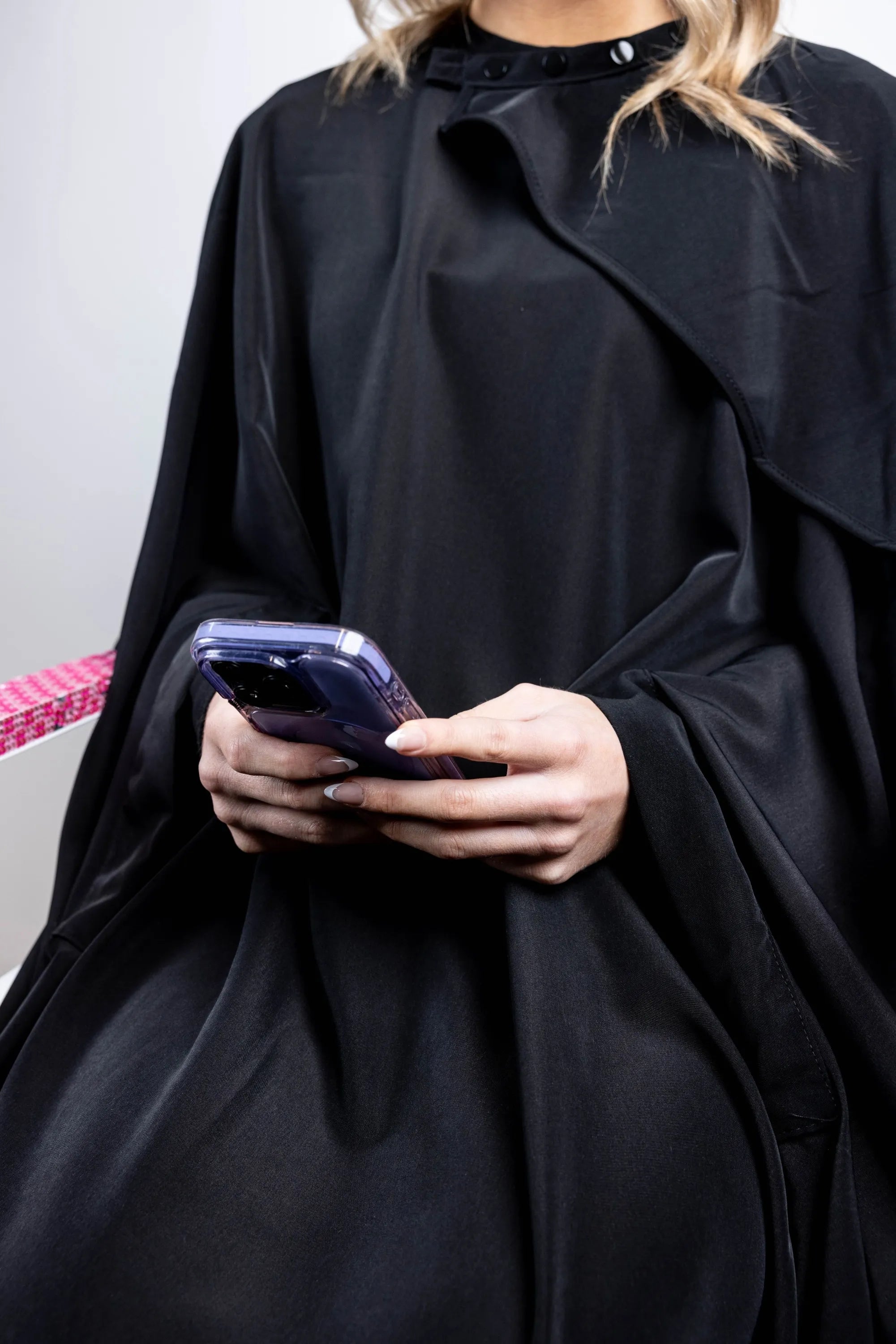 A close-up shot of a client seated in a black salon cape, holding a smartphone with both hands, highlighting the cape's snap neck closure and draped fabric.