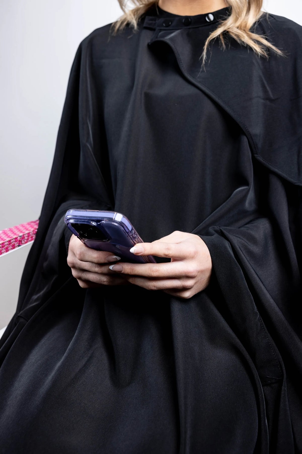 A close-up shot of a client seated in a black salon cape, holding a smartphone with both hands, highlighting the cape's snap neck closure and draped fabric.