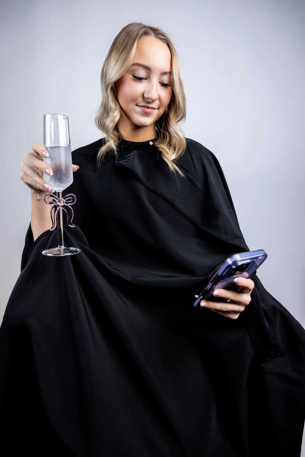 A lifestyle photograph of a woman wearing a professional black salon cape, multi-tasking by holding a champagne flute with a pink bow and looking at her smartphone while seated.