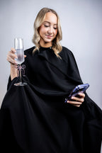 A lifestyle photograph of a woman wearing a professional black salon cape, multi-tasking by holding a champagne flute with a pink bow and looking at her smartphone while seated.