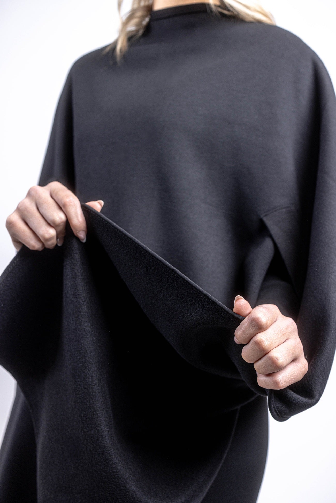 A close-up view of a person's hands lifting the fabric of a black salon cape to showcase its texture and integrated arm slit design against a plain background.