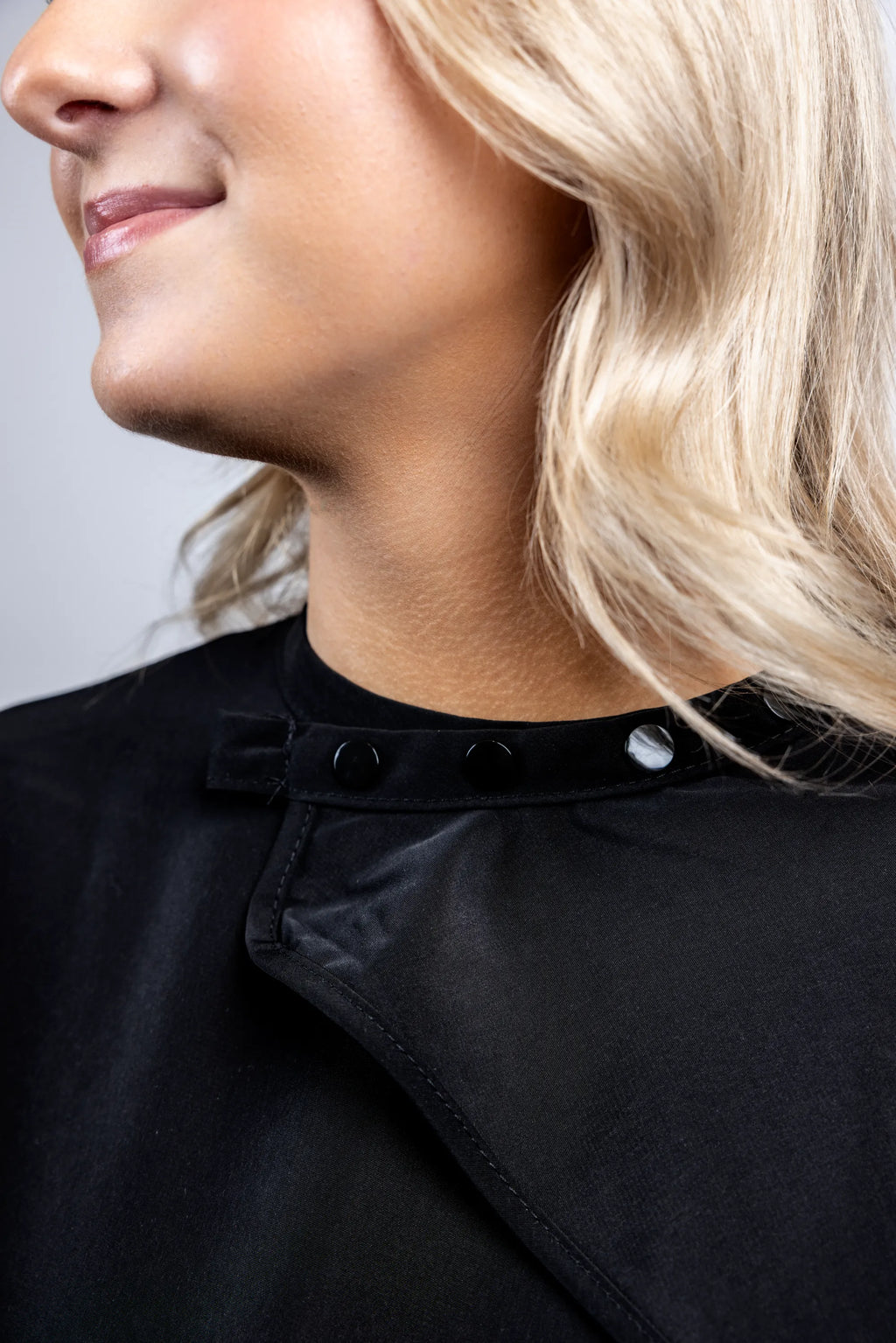 A close-up detail shot focusing on the black snap-button neck closure of a professional black salon cape, showing the secure and adjustable fasteners.