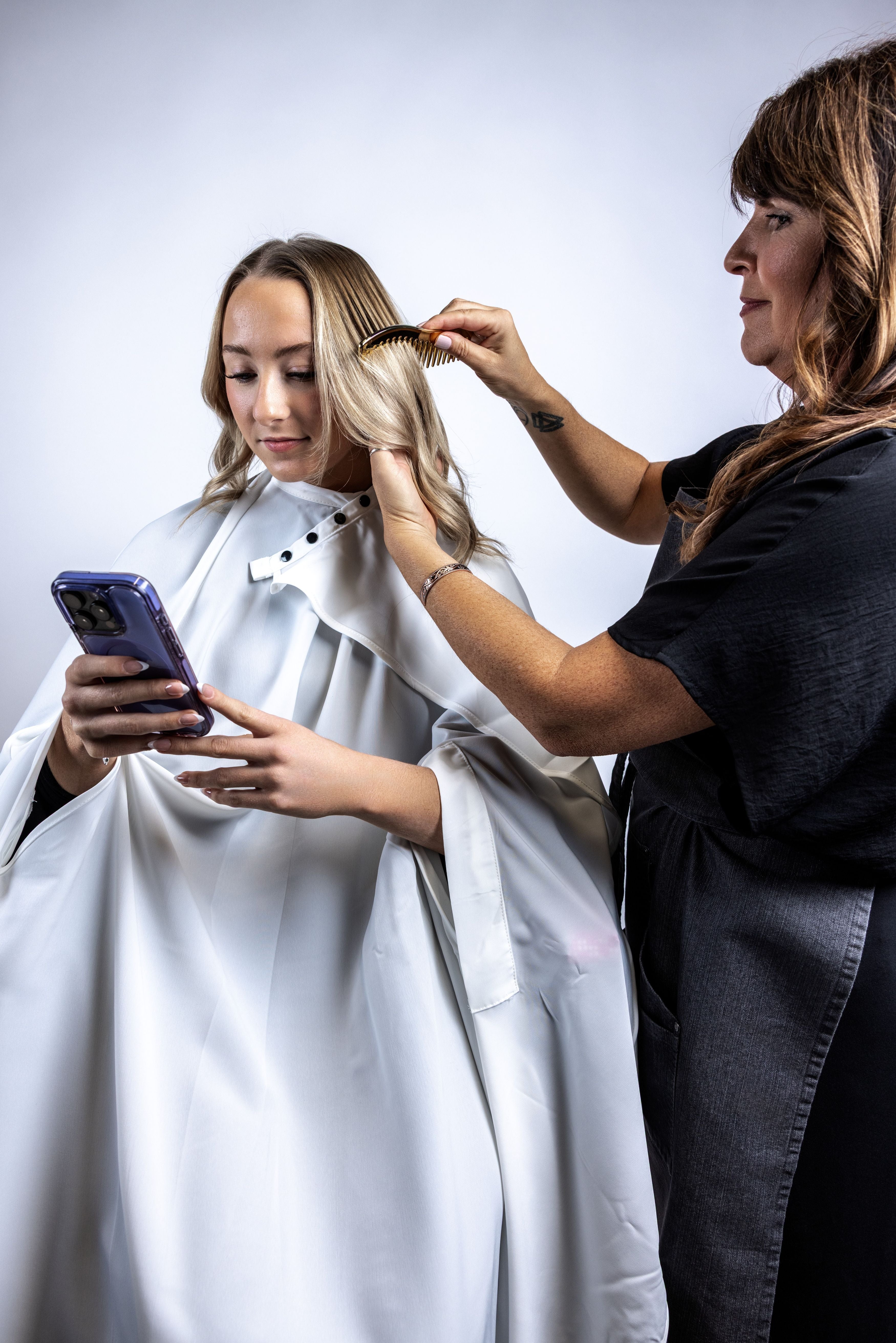 A salon lifestyle shot of a stylist combing a client's hair while she wears a white salon cape with a snap neck closure and looks at her smartphone.