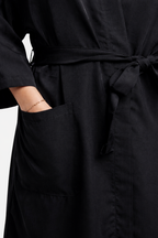 A close-up detail shot of a black salon robe or client wrap featuring a tied belt at the waist and a hand tucked into a pocket wearing a silver chain link bracelet.
