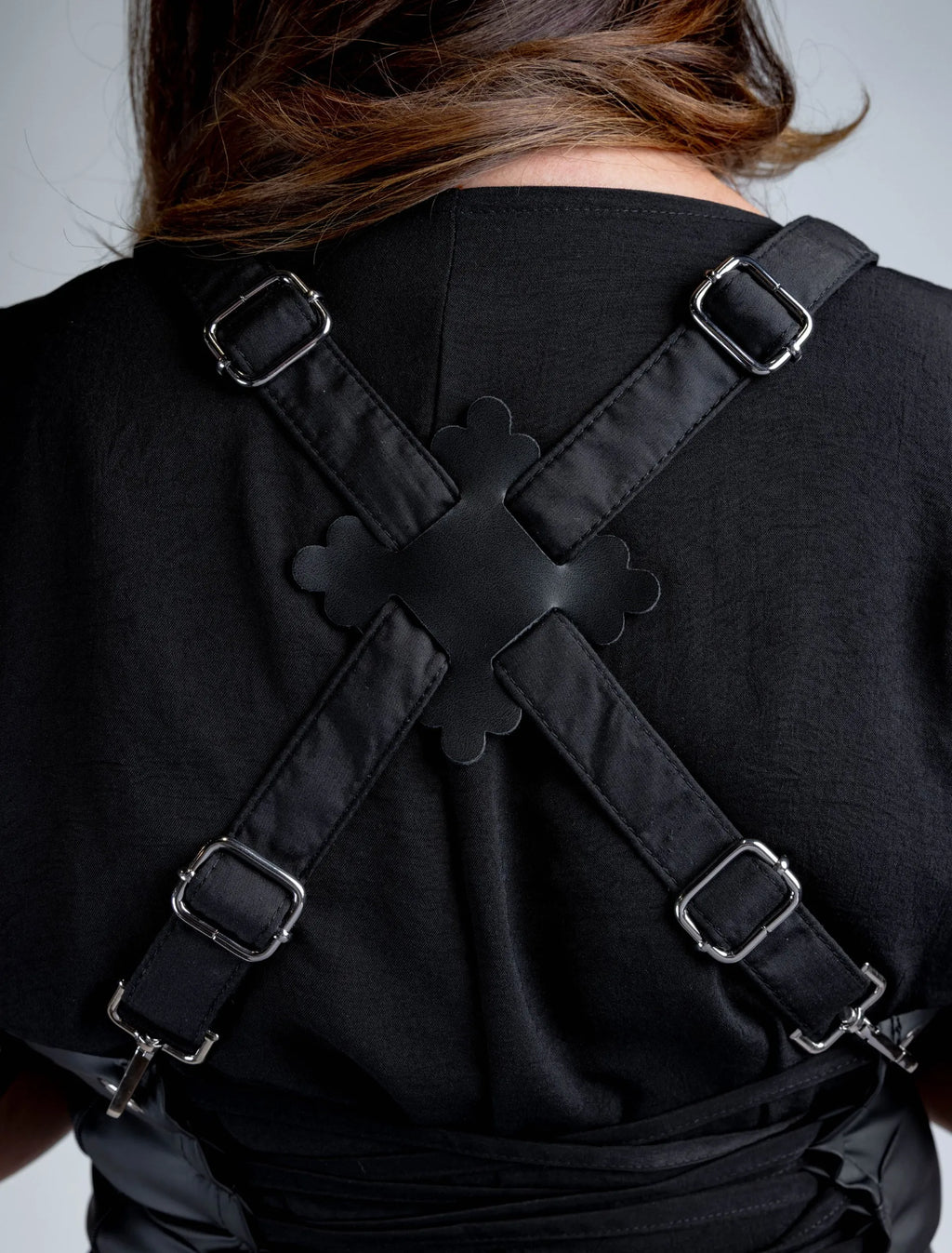 A close-up detail shot of the back of a black stylist apron, showcasing the adjustable cross-back strap system with metal hardware and a decorative central connector.