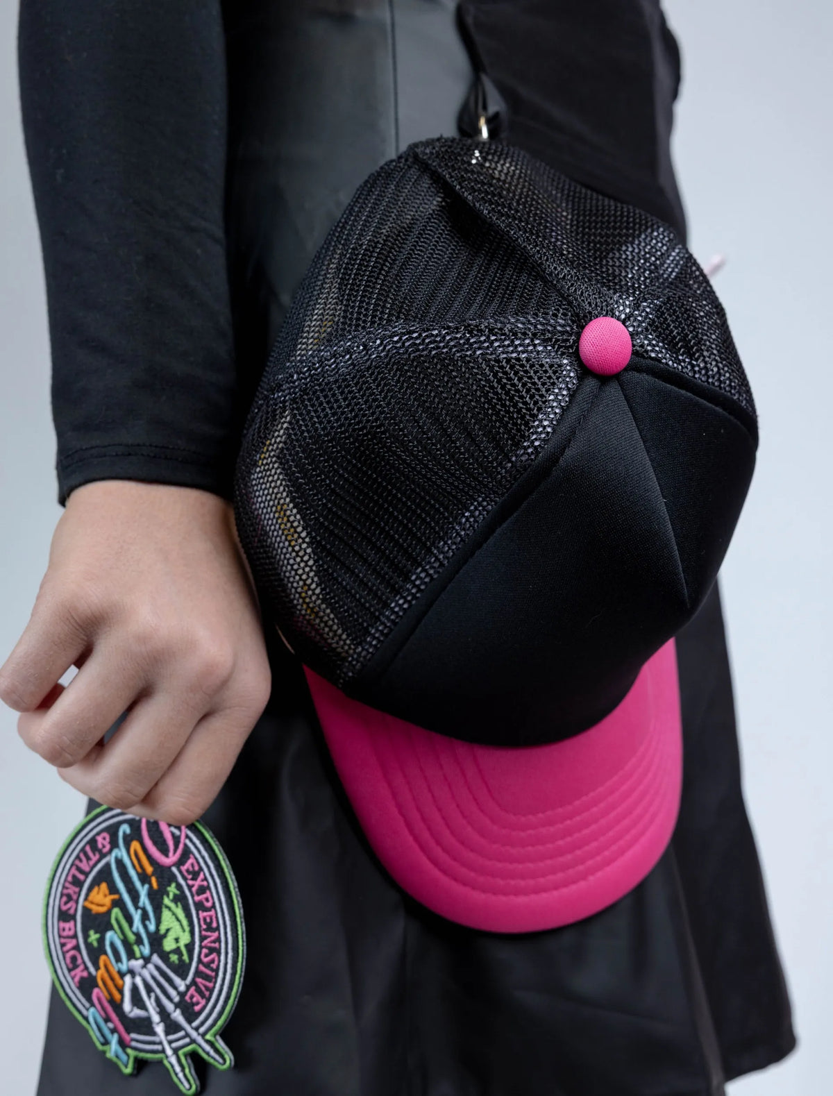 A close-up detail photograph showing a black and pink mesh trucker hat attached to a stylist apron, with a hand holding an embroidered "Expensive & Difficult & Talks Back" patch in the foreground.