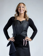 A full-length front-view photograph of a smiling blonde woman wearing a black professional stylist apron from The Branded Salon, featuring a sweetheart neckline, a central tool organizer panel with pink hair clips, professional combs, and a towel ring attachment.