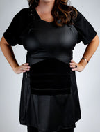 A front-view detail shot of a black stylist apron from The Branded Salon, featuring a sweetheart neckline and a central black tool organizer panel with multiple horizontal pockets and a zippered bottom section.