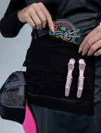 A close-up detail photograph of a black stylist apron from The Branded Salon, featuring a central tool panel with horizontal pockets holding pink hair clips and a hand placing a colorful "Expensive & Difficult" embroidered patch into a top pocket.
