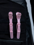 A close-up detail photograph of a black stylist apron from The Branded Salon, featuring two light pink hair clips attached to the horizontal straps of a specialized tool organizer panel.