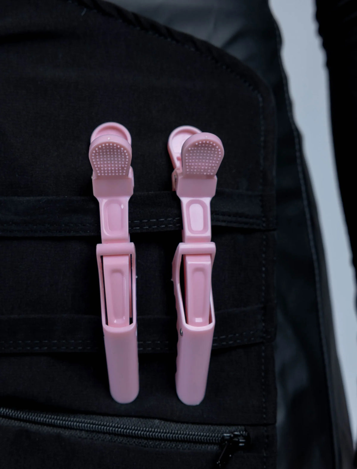 A close-up detail photograph of a black stylist apron from The Branded Salon, featuring two light pink hair clips attached to the horizontal straps of a specialized tool organizer panel.
