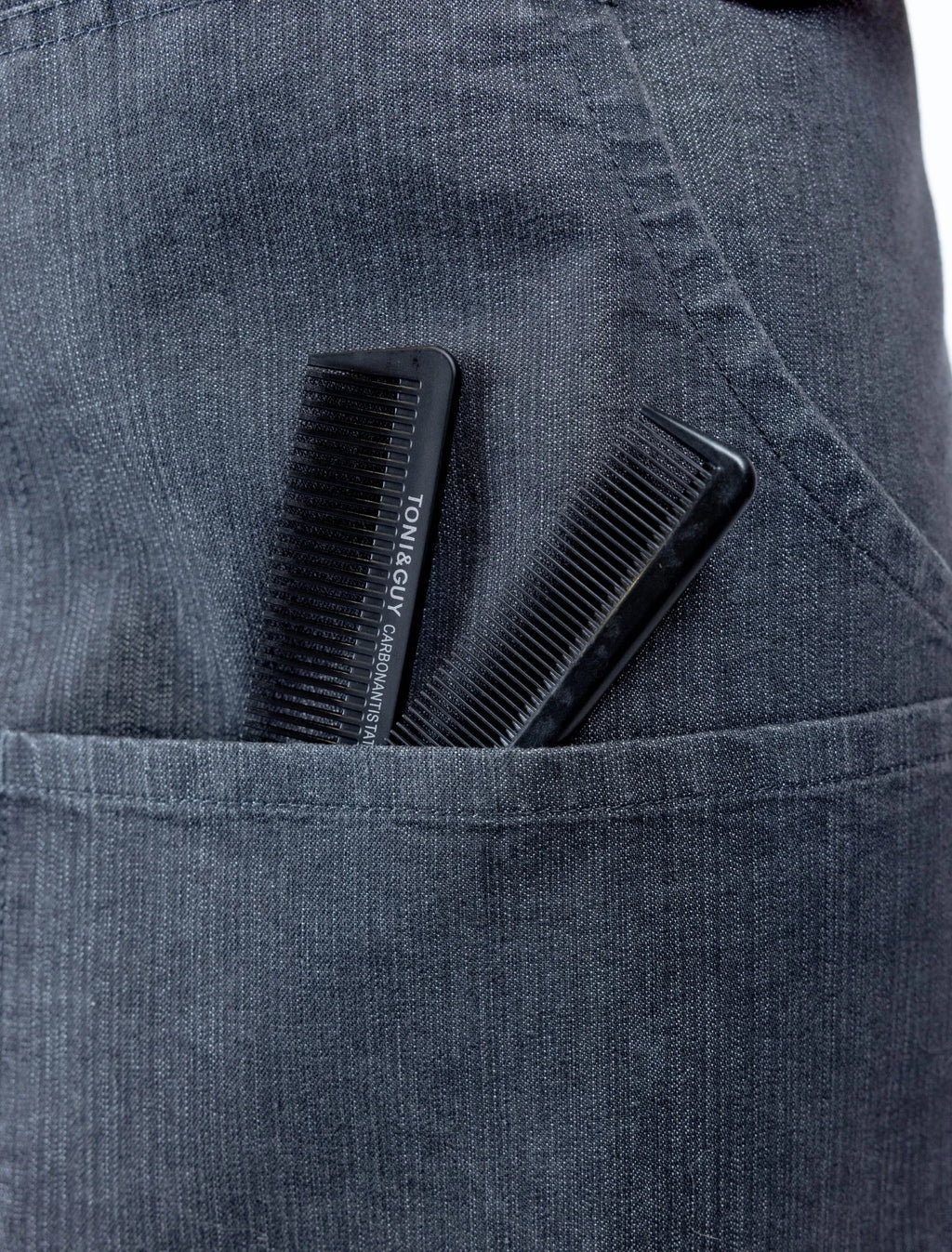 A close-up detail photograph of a charcoal grey denim stylist apron from The Branded Salon, showing two black professional combs, including a TONI&GUY carbon antistatic comb, tucked into a functional front pocket.