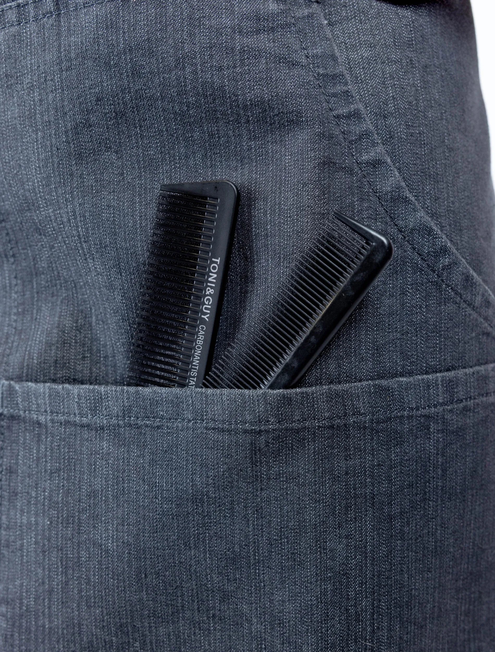 A close-up detail photograph of a charcoal grey denim stylist apron from The Branded Salon, showing two black professional combs, including a TONI&GUY carbon antistatic comb, tucked into a functional front pocket.