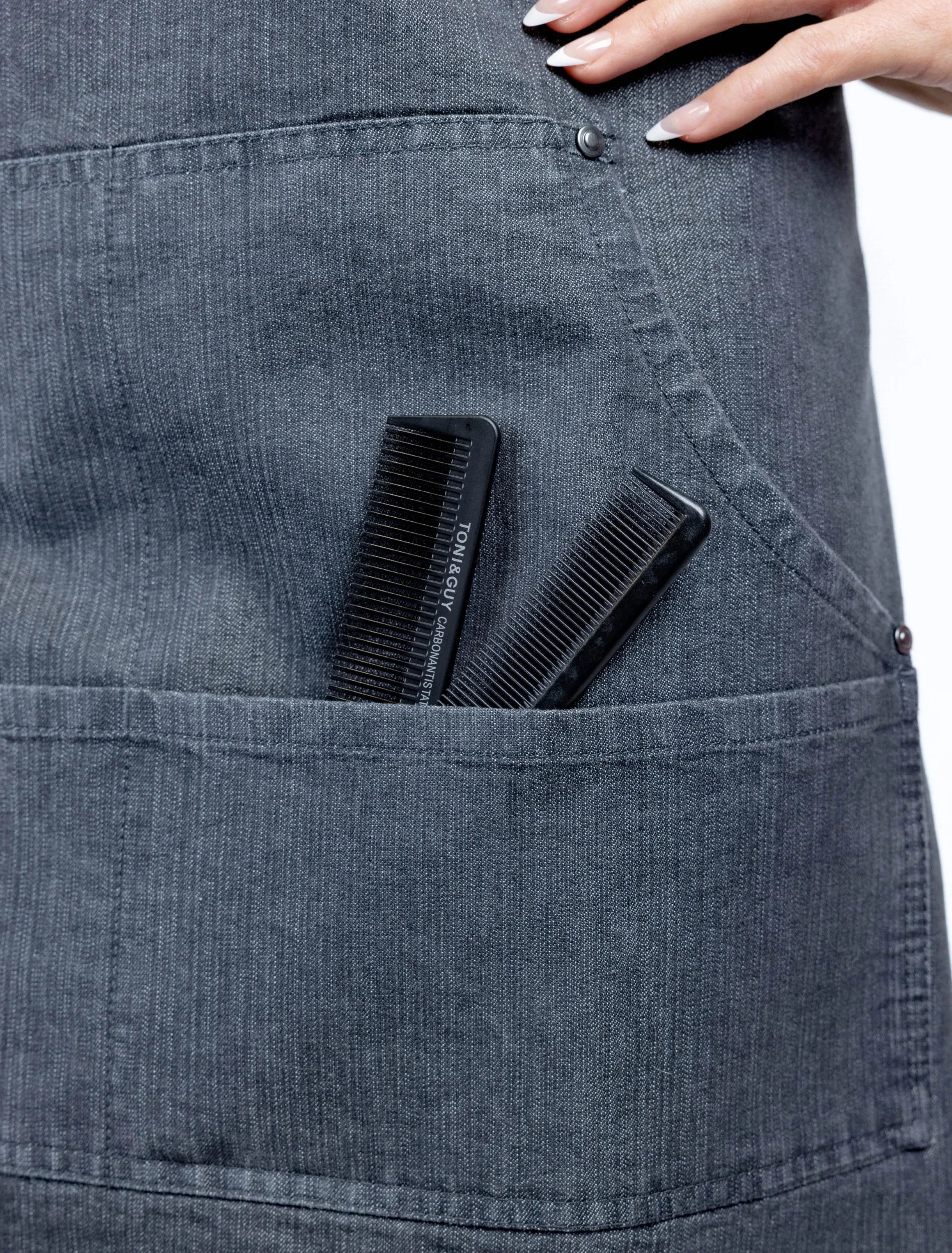 A close-up detail photograph of a charcoal grey denim stylist apron from The Branded Salon, showing two black professional combs, including a TONI&GUY carbon antistatic comb, tucked into a functional front pocket.