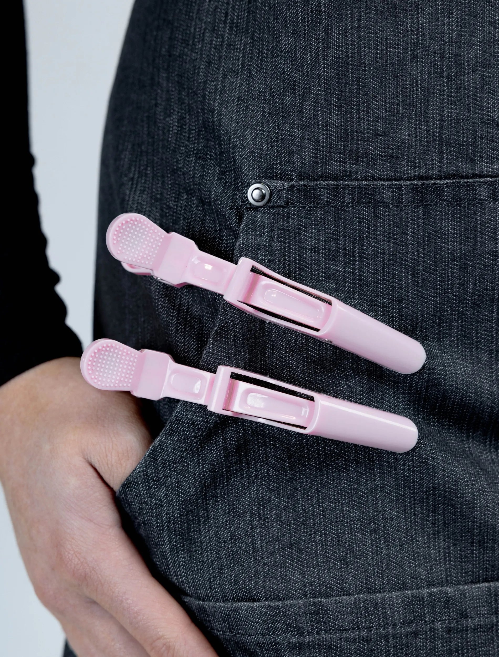 A close-up detail photograph of a charcoal grey denim stylist apron from The Branded Salon, featuring two light pink hair clips attached to the edge of a functional pocket with a hand partially tucked inside.