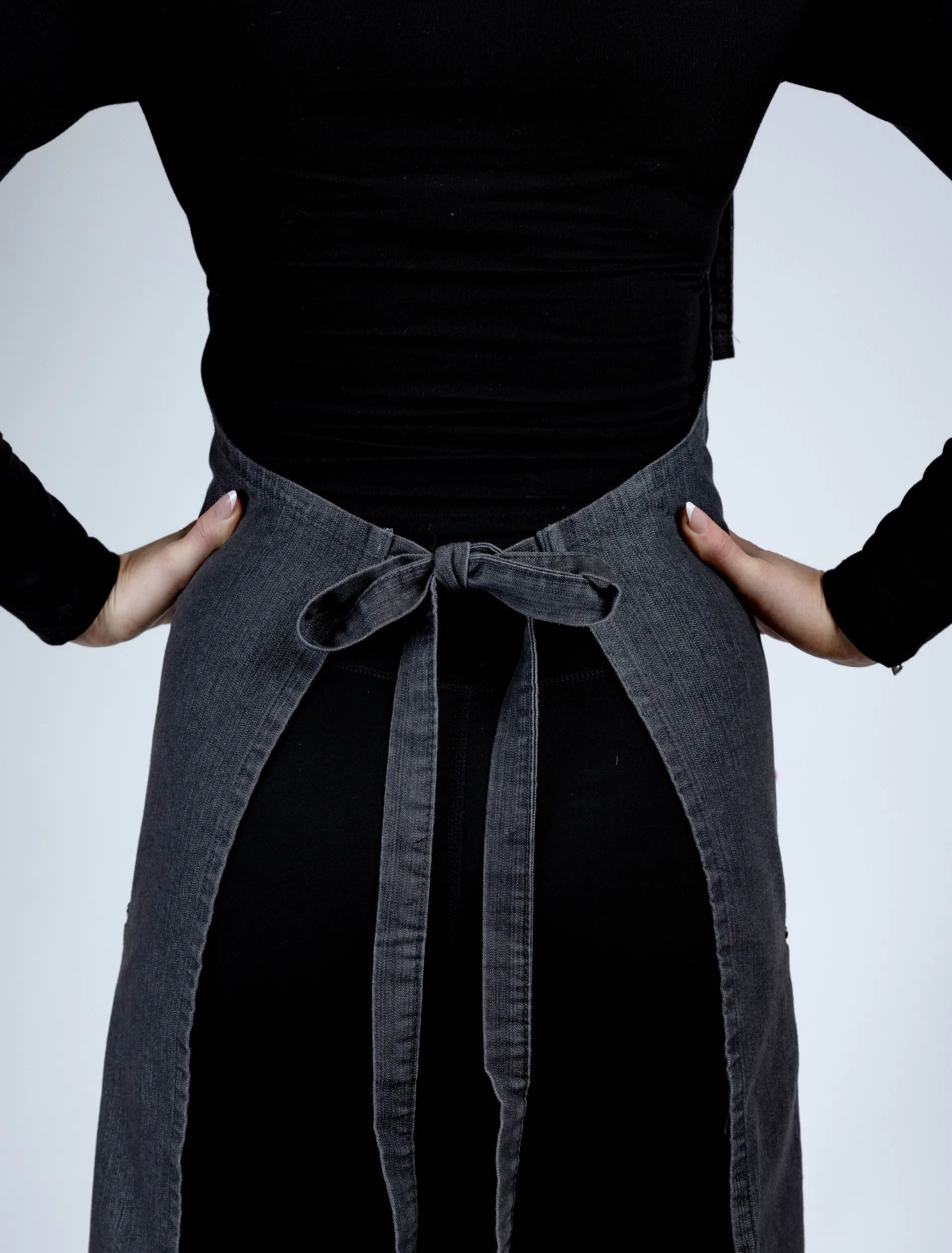 A rear-view lifestyle photograph of a charcoal grey denim stylist apron from The Branded Salon, showing the waist ties secured in a bow with the person's hands resting on their hips.