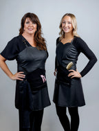A professional lifestyle portrait of two smiling stylists standing side-by-side, wearing black salon aprons with sweetheart necklines and functional tool pockets containing a gold comb and pink hair clips.