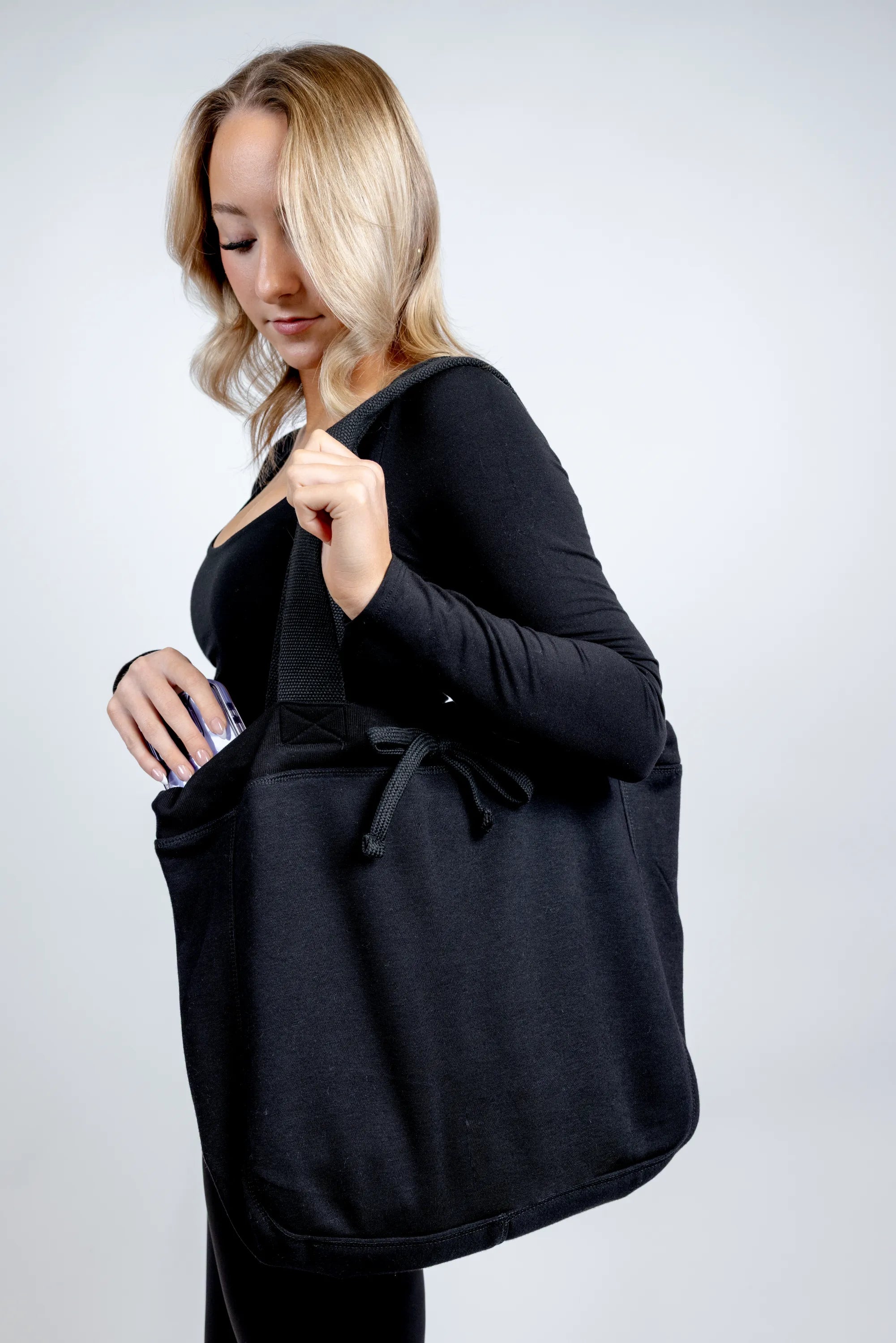 A side-view lifestyle photograph of a woman carrying a large "Black Beauty" fabric tote bag from The Branded Salon on her shoulder, reaching inside to store or retrieve her smartphone against a plain background.