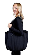 A lifestyle photograph featuring a smiling woman looking over her shoulder while carrying a large, professional black fabric tote bag from The Branded Salon on her shoulder against a plain background.