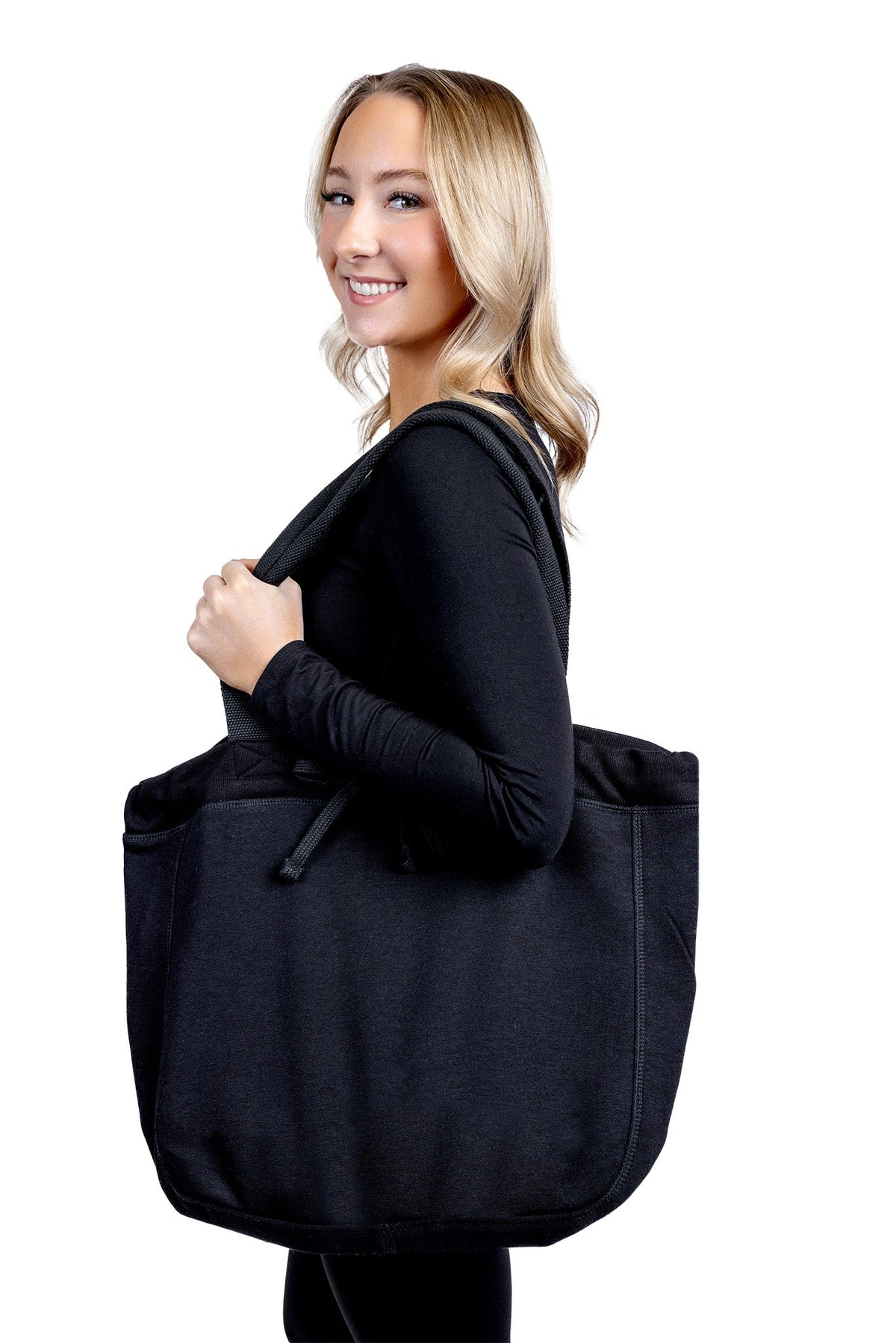 A lifestyle photograph featuring a smiling woman looking over her shoulder while carrying a large, professional black fabric tote bag from The Branded Salon on her shoulder against a plain background.