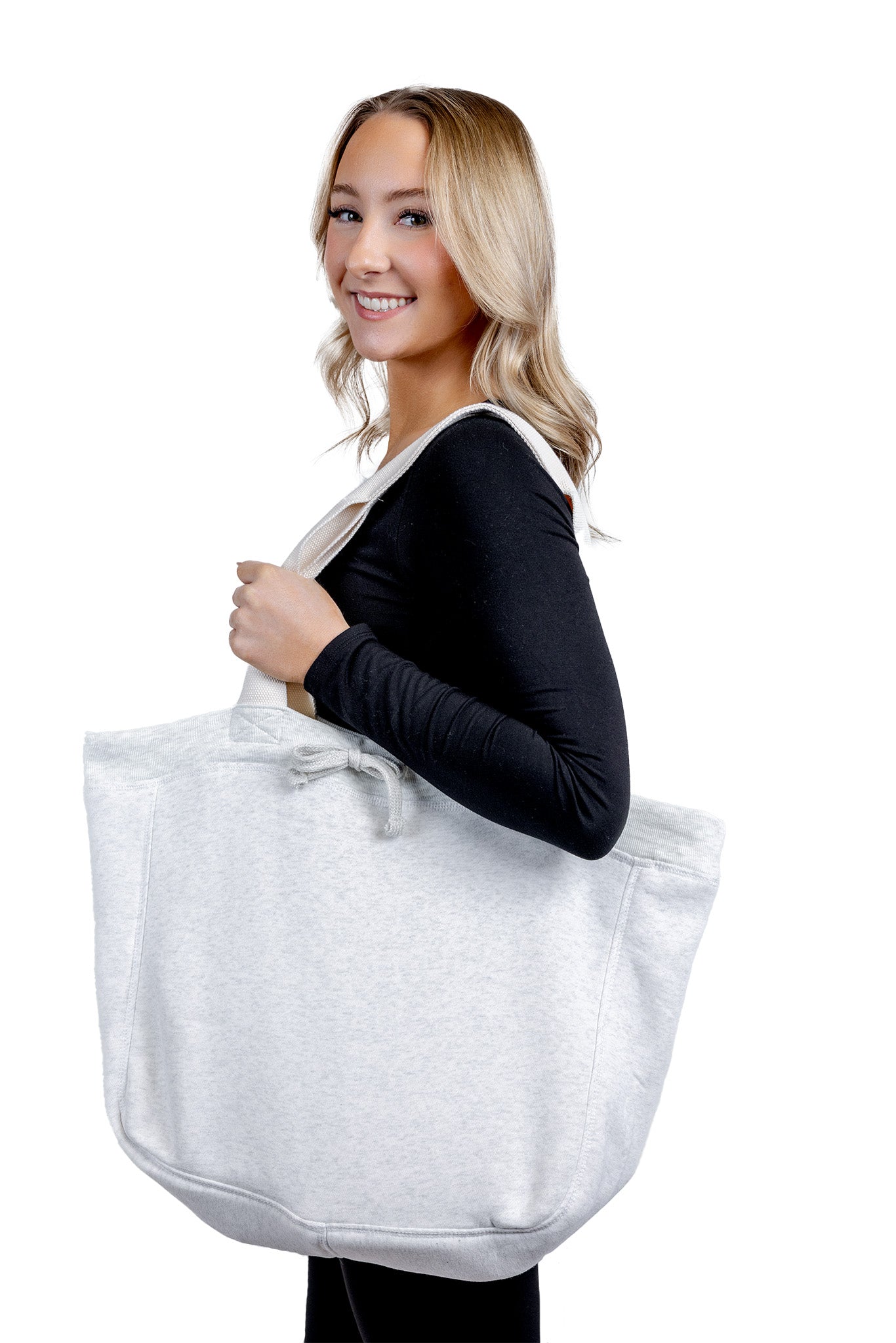 A side-view lifestyle photograph of a smiling woman carrying a large, light grey fabric tote bag with a drawstring closure detail on her shoulder against a plain background.