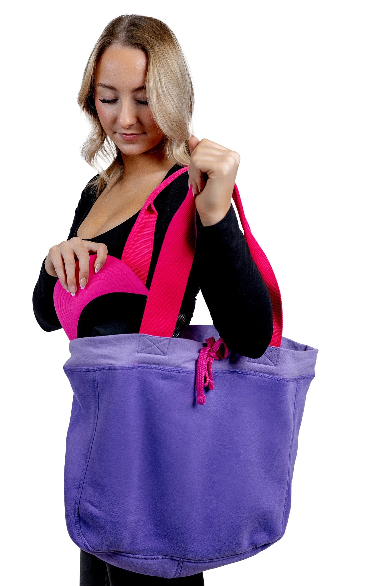 A lifestyle photograph of a woman carrying a large purple fabric tote bag with vibrant pink handles, reaching inside to retrieve a black and hot pink trucker hat.