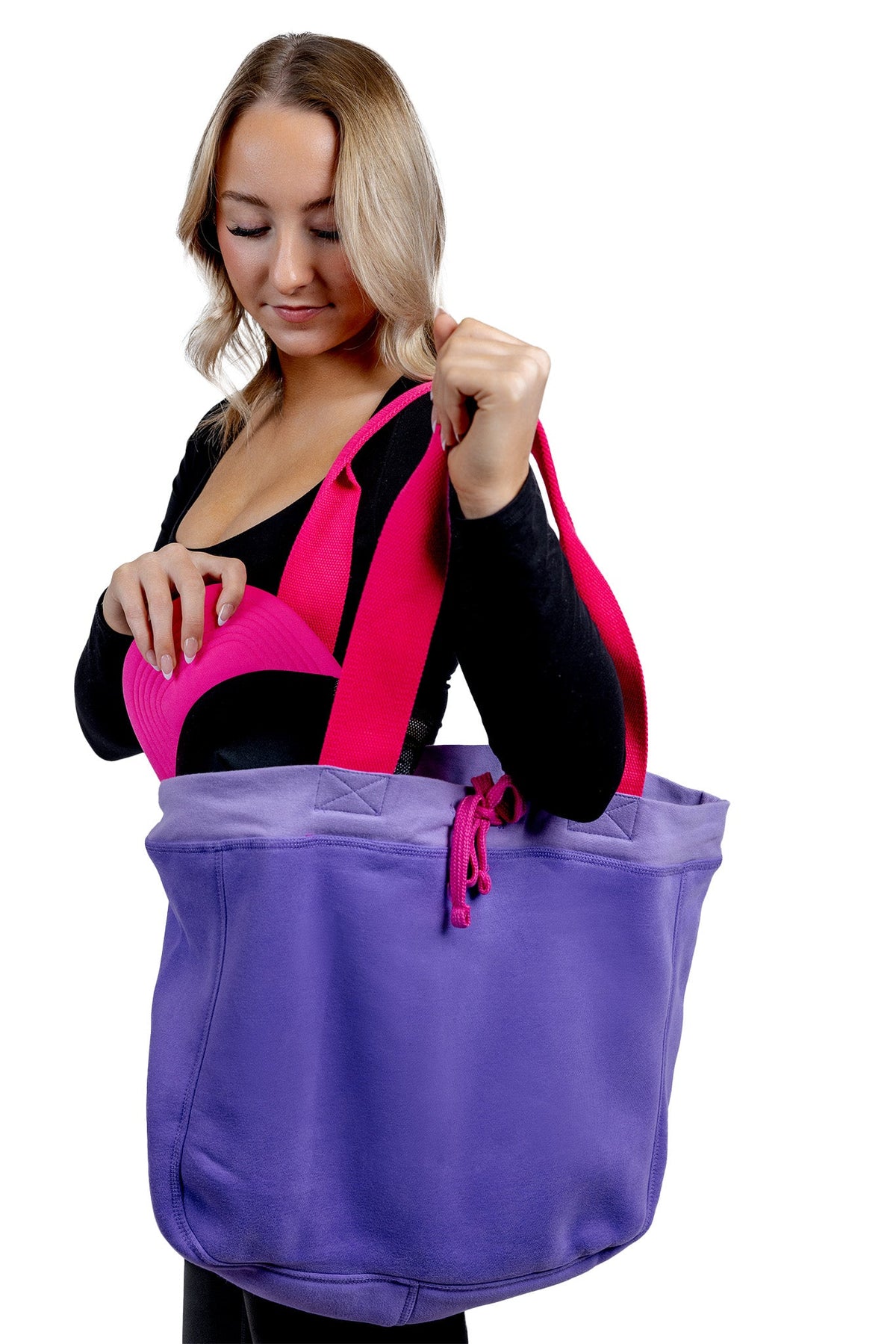 A lifestyle photograph of a woman carrying a large purple fabric tote bag with vibrant pink handles, reaching inside to retrieve a black and hot pink trucker hat.